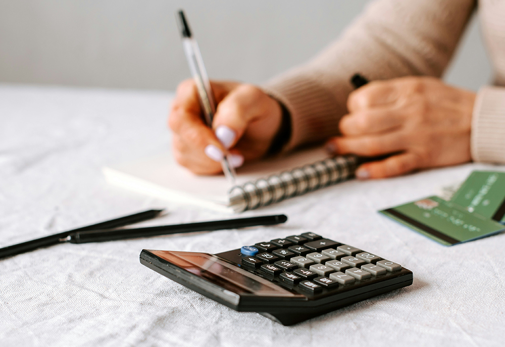 Person sat at a table with a calculator in the foreground. The person is making notes in a spiral bound notebook with credit cards on the table.
