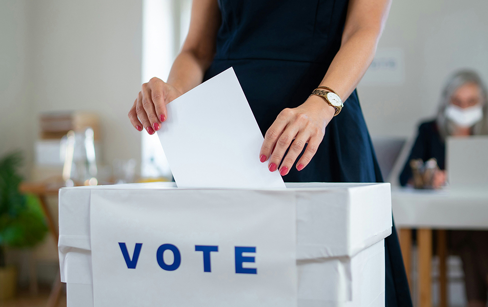 Lady in a black outfit placing a piece of paper into a box with a sign saying Vote