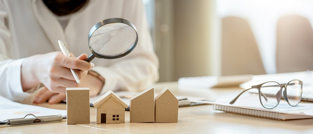 Person sat desk with magnifying glass looking at small wooden building blocks on the desk. The second block in is of a house.