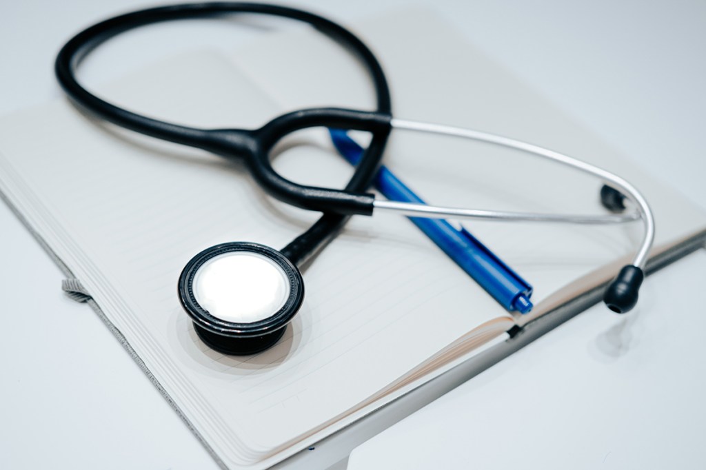 A stethoscope on top of an open notebook with a blue biro resting in the notebook's spine