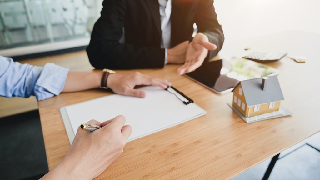 Two people sat at a table. One has a clipboard and pen. A model house is on the table.