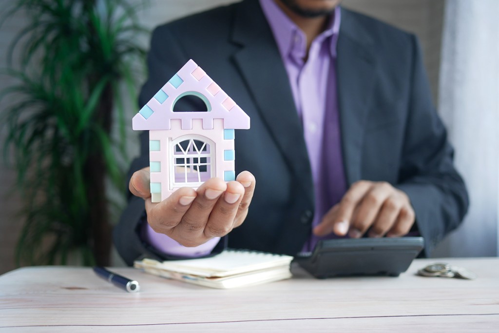 A man in a suit is using a calculator with one hand whilst there is a model house in the palm of his other hand.