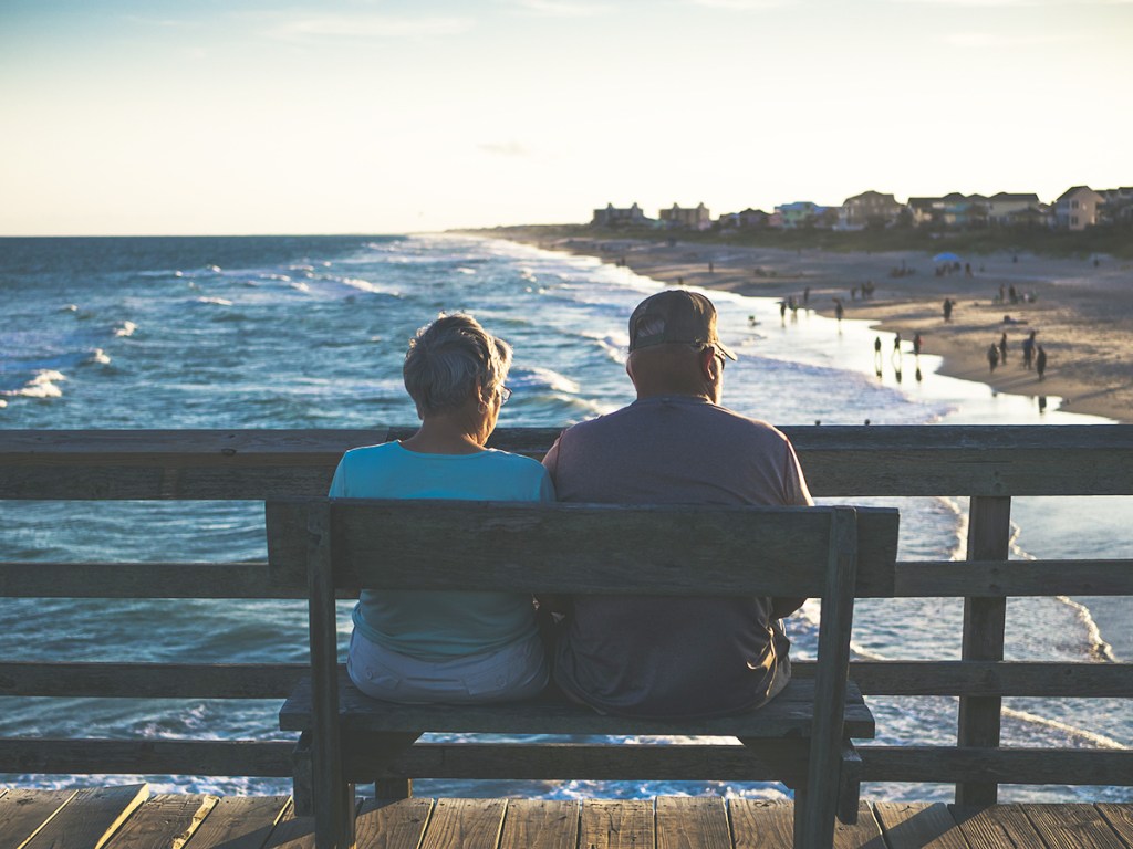 Senior couple sitting on a bench with their backs to camera, looking out over a beach with waves coming in