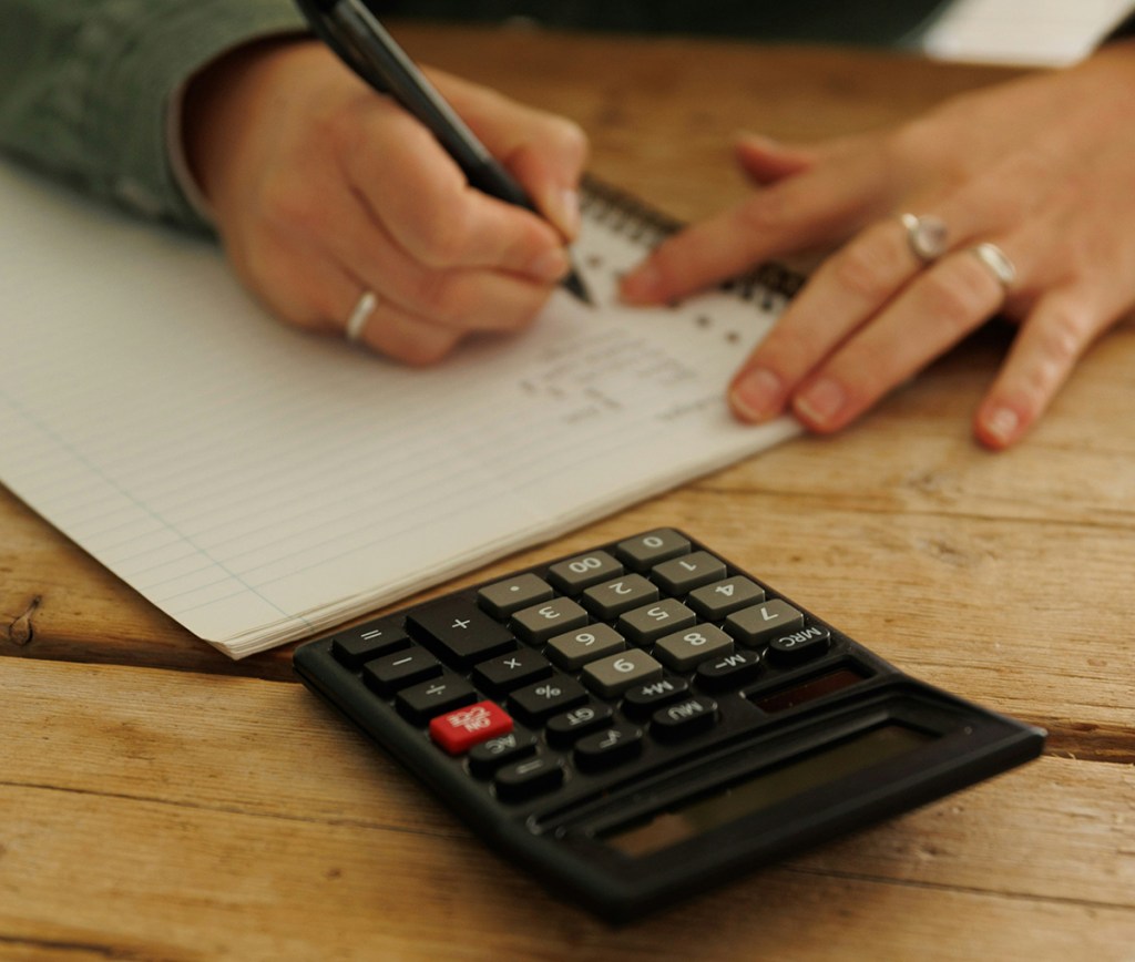 Person sat at a table writing in a notebook with a calculator above the notebook