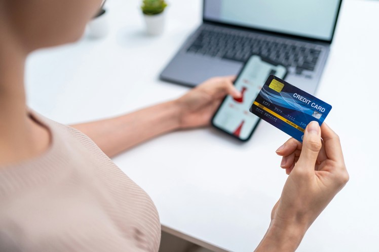 Woman holding credit card. Blurred in the background is a mobile phone with a shopping site on, and a laptop