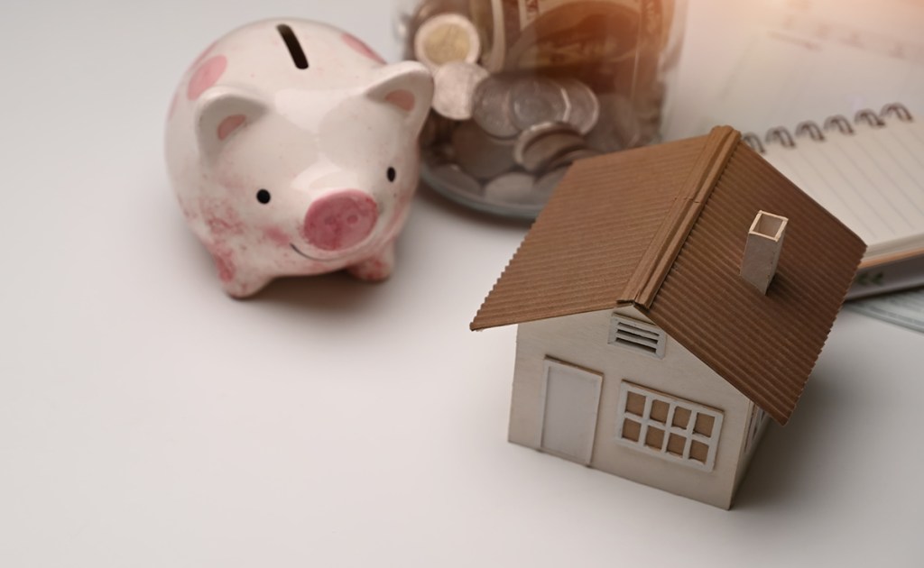 Model of a home on a table with a spiral-bound notebook behind it. A piggy bank and jar of coins is behind the model of the house