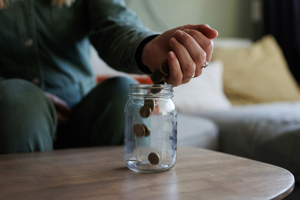 A person sat at a coffee table pour coins from the palm of their hand into a jar