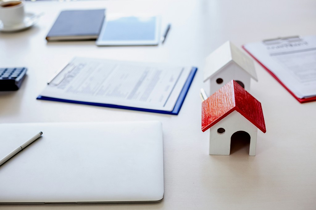 Two model homes on a table, one with a red roof and the other with a white roof. Around the models are two clipboards and a closed laptop