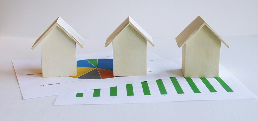 Three wooden models of houses on a table, on top of two pieces of paper with graphs on them