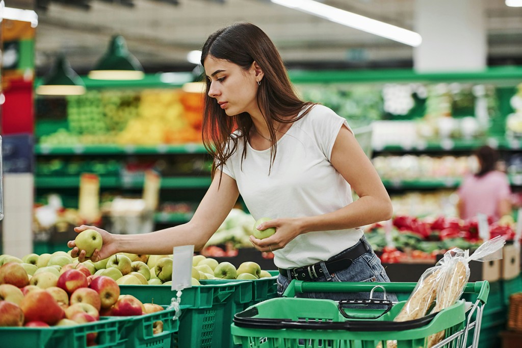 A lady in a supermarket with a green trolly standing next to apple variants, picking up a green apple.