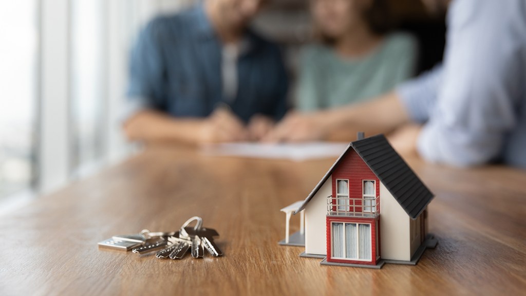 A model of a home on a table with a set of keys next to it. Out of focus in the background are three people looking at a piece of paper