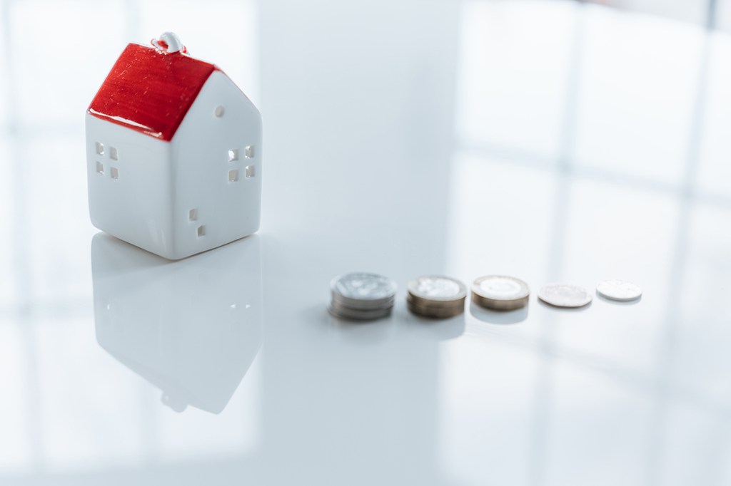 A white ceramic model of a house with a red roof on a white table. Five piles of coins decreasing in height from left to right are next to it.