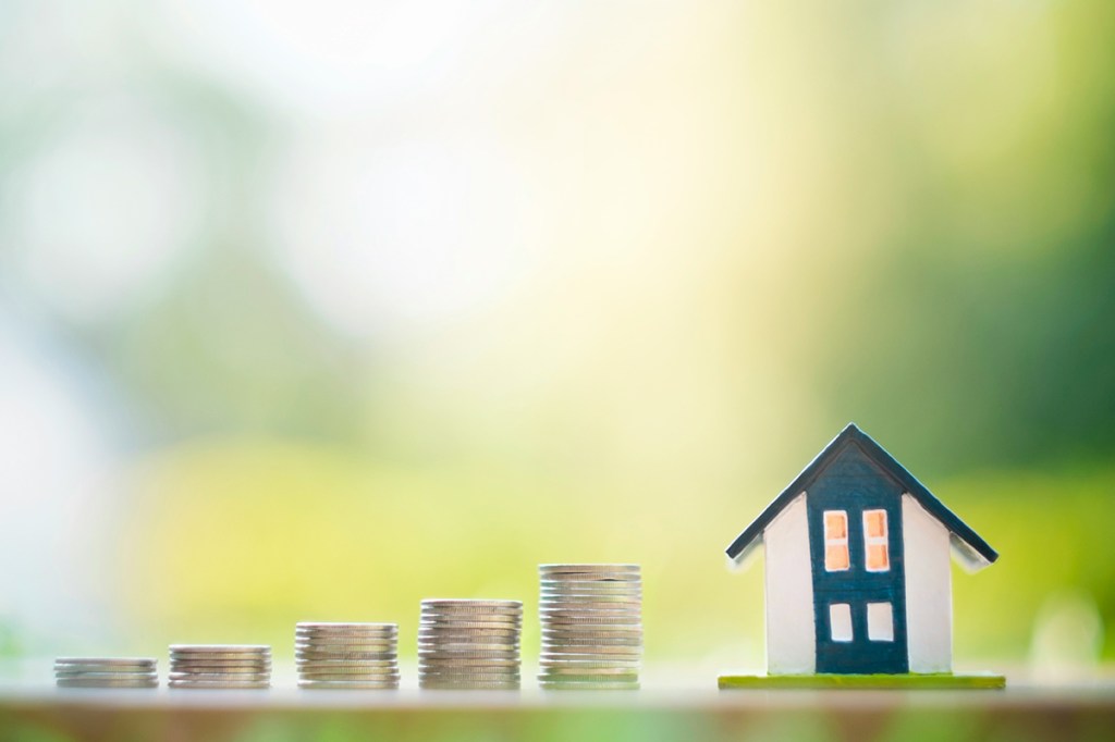 A model of a house on a table, with piles of coins to its left, increasing in size. There is a green background blurred out