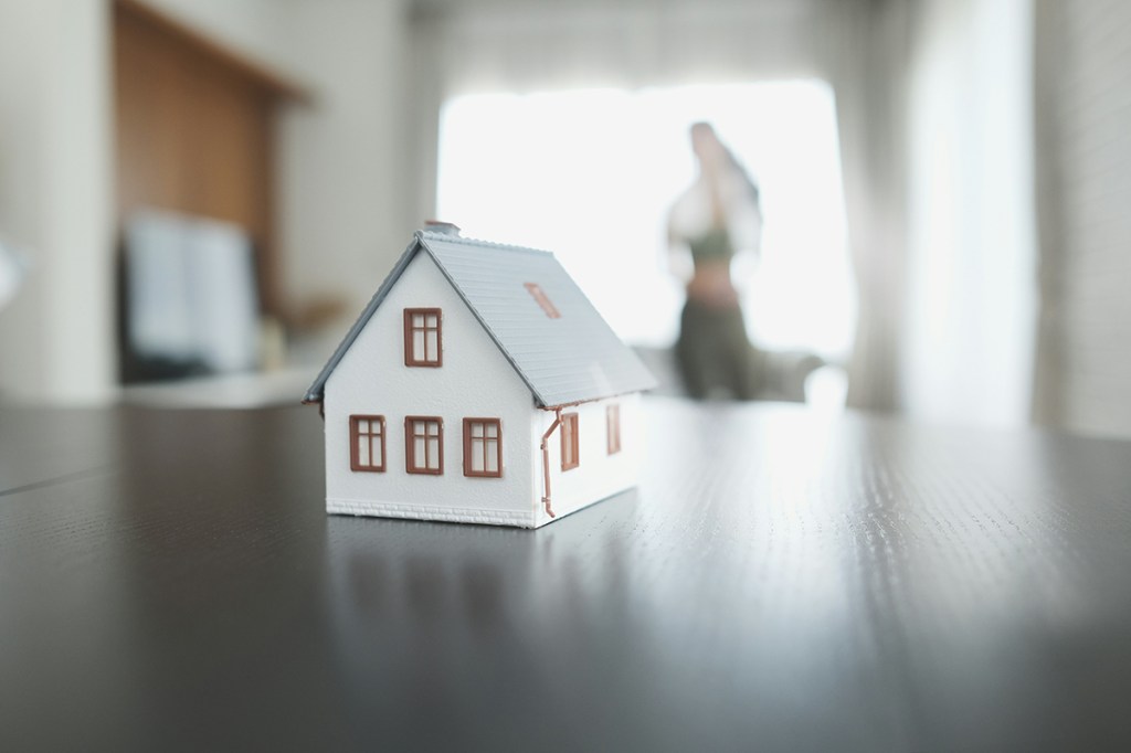 A model of a home with white walls and a grey roof and brown window frames, on a table. The background of the image is blurred