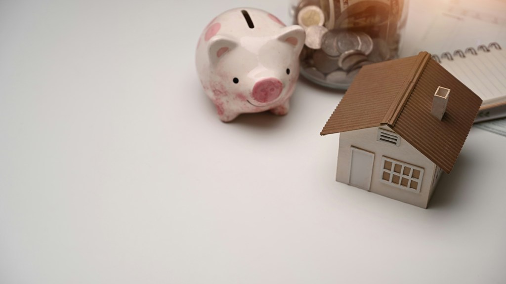 A model of a house on a table with a pink piggy bank and a jar of coins with an open spiral-bound notebook, all in the top-right of the image