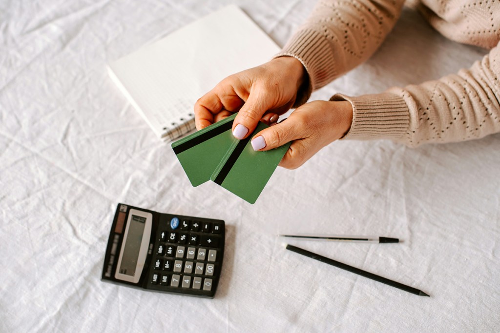 Arms leaning on a table with two green credit cards in hand. A calculator, notepad and two pens are on the table.