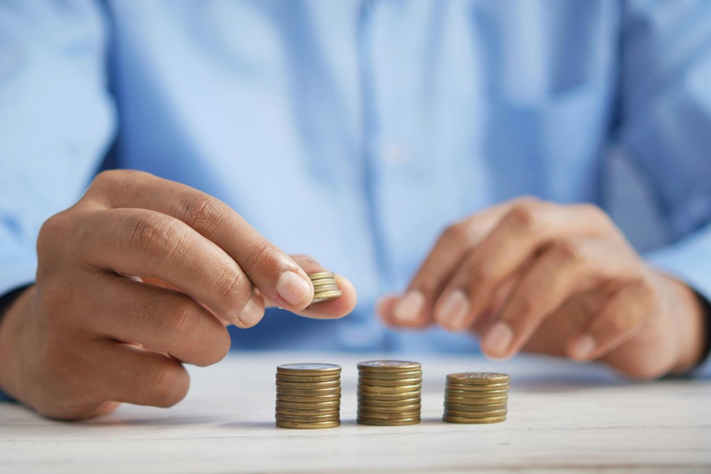 A person in a blue shirt sat at a table, arms in view. Three piles of coins are on a table. More coins are being added to the first pile.