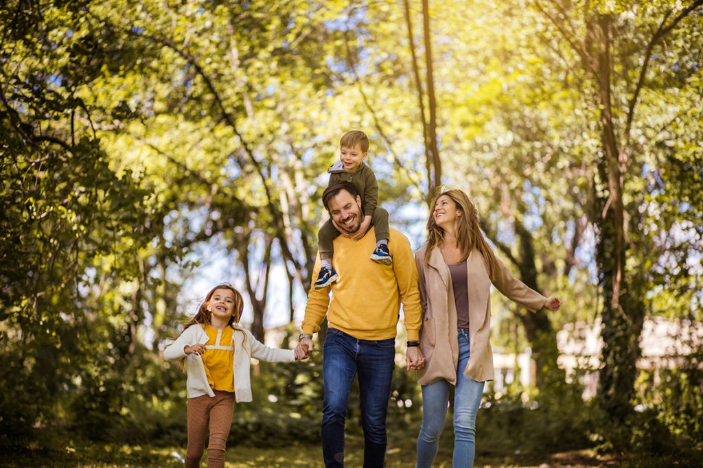 A family of two parents and two children walking through the woods. The dad has one child on his shoulders and is holding hands with the other child