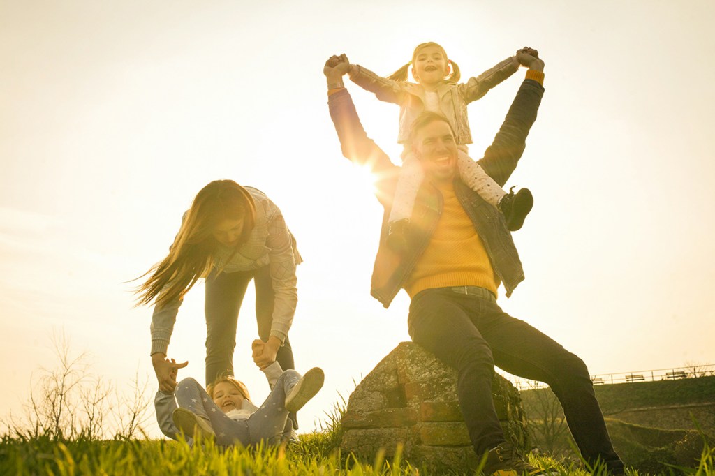 A family picture with the mum holding the hands of one child and the dad with another child on his shoulders with their arms in the air