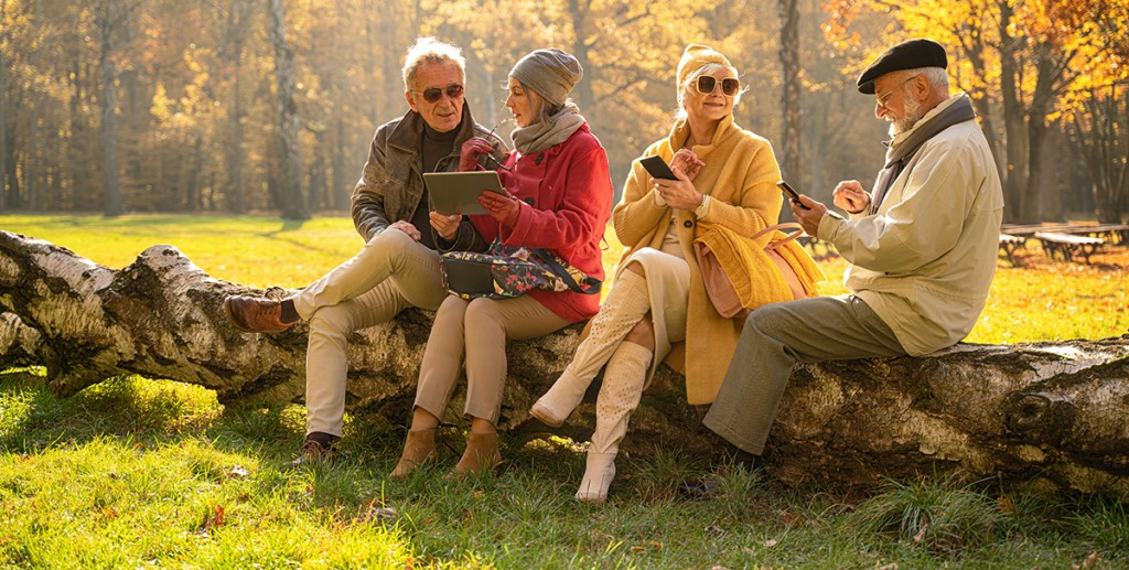 Four older people sat on a log bench in a park setting with phones and tablets in their hands