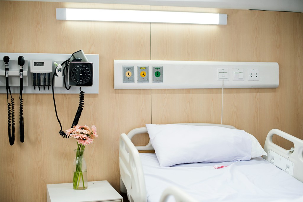 A hospital bed without a patient in, with a vase of pink daisies on the bedside table