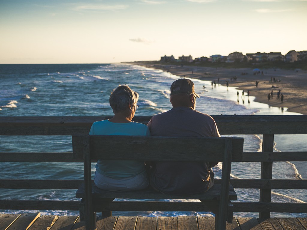 An older couple sat on a beach on a pier overlooking the sea and a beach with houses in the background